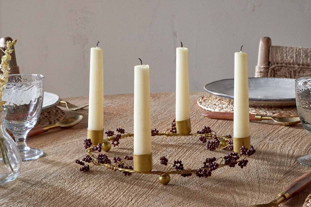 The nkuku Tanu Mango Wood Beaded Candle Holder in Brass & Ruby, adorned with small purple berries and cream candles, adds festive charm to a wooden dining table set with woven placemats, clear glasses, plates, and cutlery.