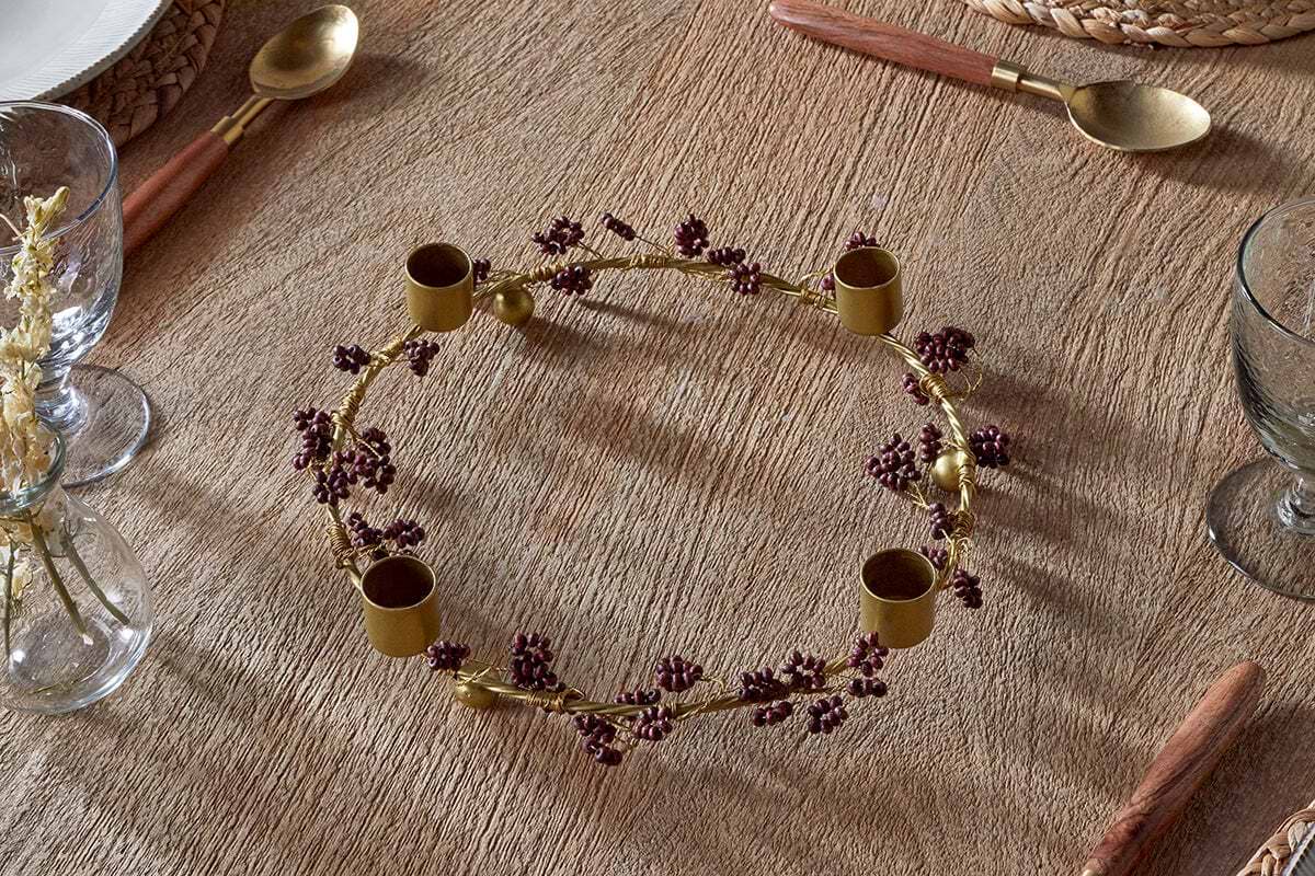 The Tanu Mango Wood Beaded Candle Holder in Brass & Ruby by nkuku adds a festive, rustic accent to a textured wooden table, surrounded by glassware and gold cutlery for a charming seasonal tablescape.