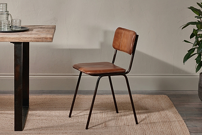 The Ukari Leather Dining Chair - Aged Tan by nkuku, featuring a padded leather seat and metal frame, sits beside a wooden table on a beige woven rug. Neutral tones and a visible plant add to the space's modern, utilitarian style.