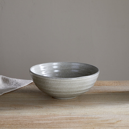 The Valinha Earthenware Cereal Bowl - Grey by nkuku features a light gray finish and textured spiral design. Shown on a wooden surface beside a beige cloth and matching pitcher, it’s set against a plain light-colored wall.