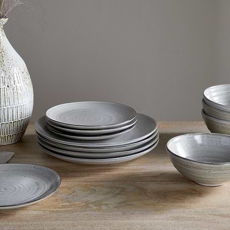 The nkuku Valinha Earthenware Dinnerware Set - Grey (Set of 12) features ceramic plates and bowls with a gray speckled glaze, arranged on a wooden table beside a cream pitcher and a folded napkin.