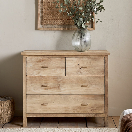 The Vivan Grooved Chest of Drawers - Natural by nkuku stands against a beige wall, topped with a clear textured vase holding greenery. An armchair with a striped cushion and woven basket sits to the left; part of a bed is visible on the right.
