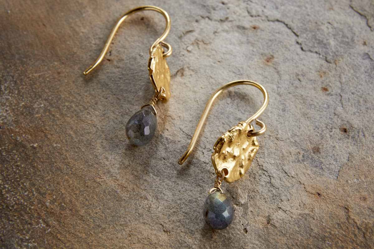 The Veera Labradorite Earrings – Gold by nkuku feature gold-coloured hooks with textured oval details and faceted labradorite drops, shown on a stone surface.