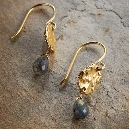 The Veera Labradorite Earrings – Gold by nkuku feature gold-coloured hooks with textured oval details and faceted labradorite drops, shown on a stone surface.