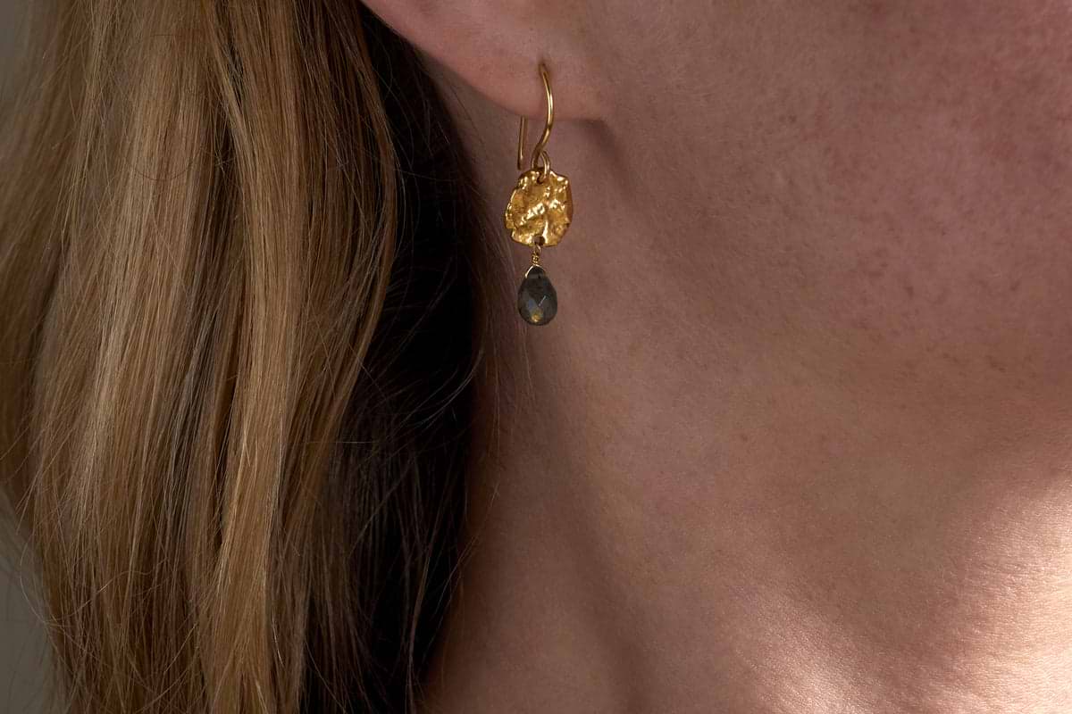 Close-up of a womans ear wearing nkukus Veera Labradorite Earrings - Gold, featuring a textured round gold top and a dark teardrop-shaped labradorite stone. Light brown hair is partially visible.