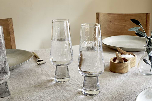 Two clear Visla Champagne Flutes by nkuku are filled and placed on a beige tablecloth. The table features light gray plates, wooden chairs, a fork, a small wooden salt cellar, and a vase with green leaves.