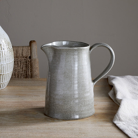 The Valinha Earthenware Jug in grey by nkuku rests on a wooden table beside a folded beige cloth, with a textured vase and ceramic cup in the background, creating a calm, rustic ambiance perfect for unique dining.