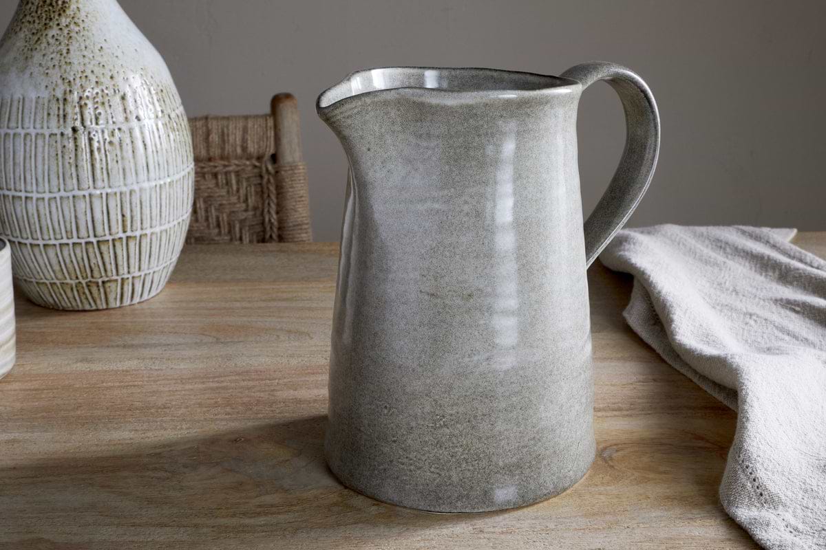 The Valinha Earthenware Jug in Grey (Large) from nkuku sits on a wooden table, bringing elegance to your serveware. Behind it, a textured vase and woven chair enhance the earthy decor, while a draped cloth softens the dining scene.