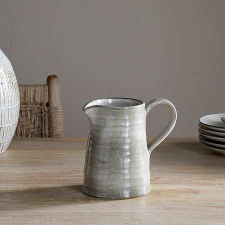 The Valinha Earthenware Jug - Grey - Small by nkuku rests on a wooden table, accompanied by stacked plates, a cloth, and a large textured vase. A woven chair completes this calm, rustic setting—ideal for an elevated dining experience.