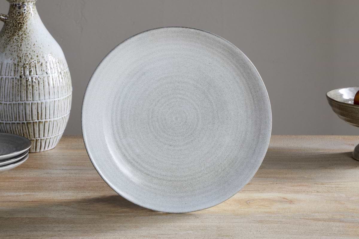 The Valinha Earthenware Dinnerware Set - Grey by nkuku, featuring a round, light gray ceramic plate with a subtle textured pattern and reactive glaze, is shown upright on a wooden table among other serveware and pottery.