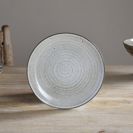The Valinha Earthenware Side Plate - Grey by nkuku, featuring a light gray reactive glaze and subtle spiral texture, stands upright on a wooden table with stacked plates and a woven chair to the left and a bowl of red apples to the right.