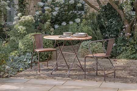 The nkuku Vivika Outdoor Dining Table - Natural sits on a stone patio, paired with two matching metal chairs with wooden seats. Stacked plates and napkins rest on the table, while lush greenery and white flowers fill the garden backdrop.