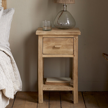 The Vivan Grooved Bedside Table - Natural by nkuku holds a glass lamp, drinking glass, and two books on its lower shelf beside a bed with neutral bedding and a woven basket on the wooden floor.
