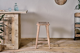 The Vinay Woven Counter Stool - Natural by nkuku, with a jute seat and acacia wood frame, is placed at a rustic wooden bar in a room with plank flooring and a textured wall finish.