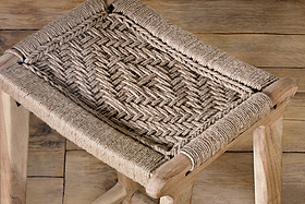 A close-up of the Vinay Woven Counter Stool by nkuku, featuring a natural jute seat in a herringbone pattern and four sturdy acacia wood legs, set on a weathered wooden floor.