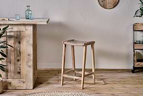A rustic interior showcases the nkuku Vinay Woven Counter Stool – Natural at a wooden bar counter, complemented by a small metal plant shelf on the right and a round woven rug partially seen on the wooden floor.