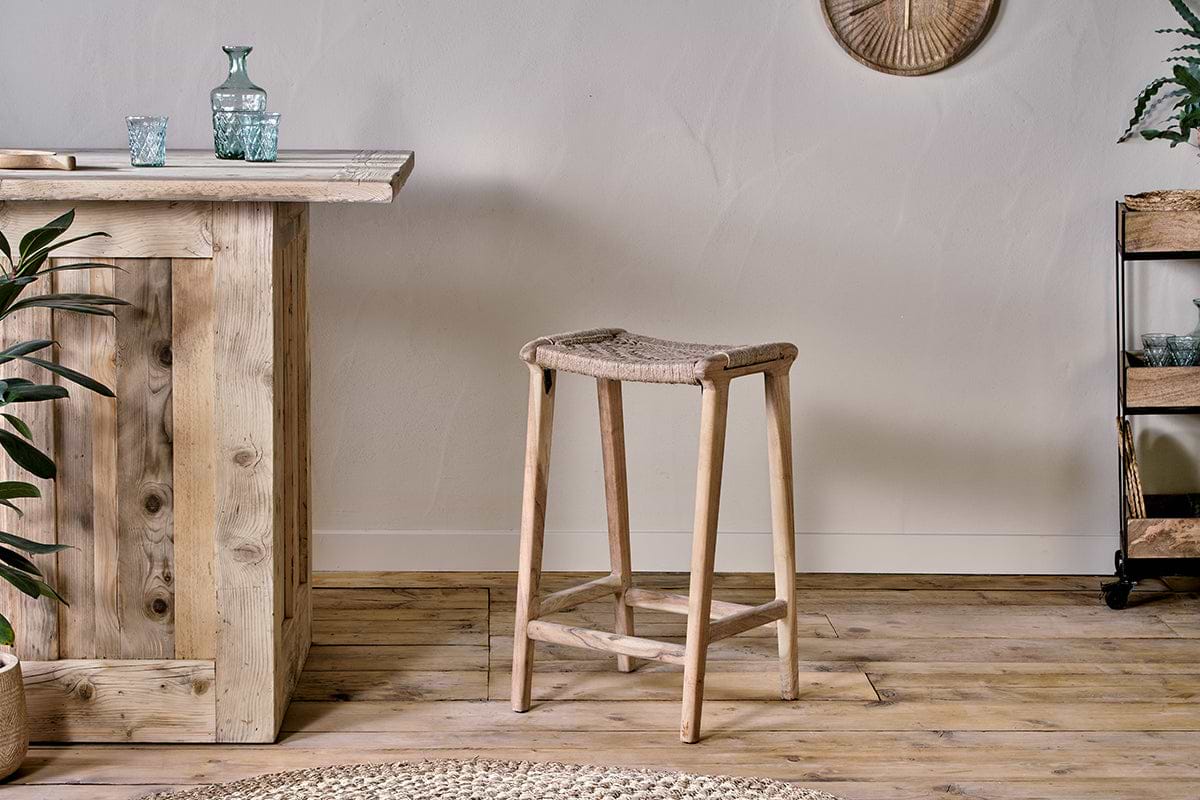 A rustic interior showcases the nkuku Vinay Woven Counter Stool – Natural at a wooden bar counter, complemented by a small metal plant shelf on the right and a round woven rug partially seen on the wooden floor.