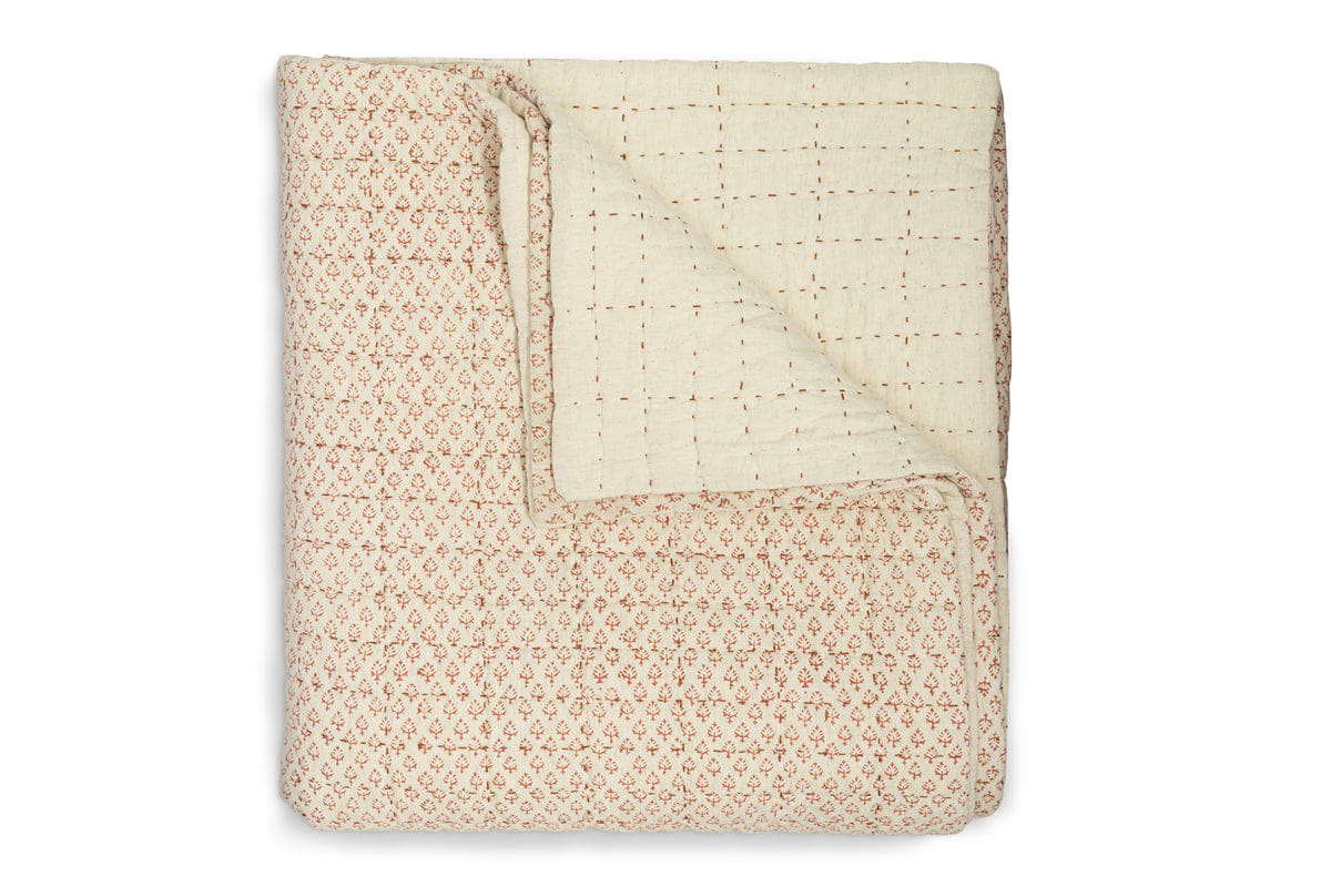The Peplu Cotton & Linen Screen Print Bed Quilt - Rust by nkuku is a folded, handcrafted piece with one corner turned over, displaying a red floral pattern on one side and a cream stitched grid on the other—beautifully crafted Indian textile art.