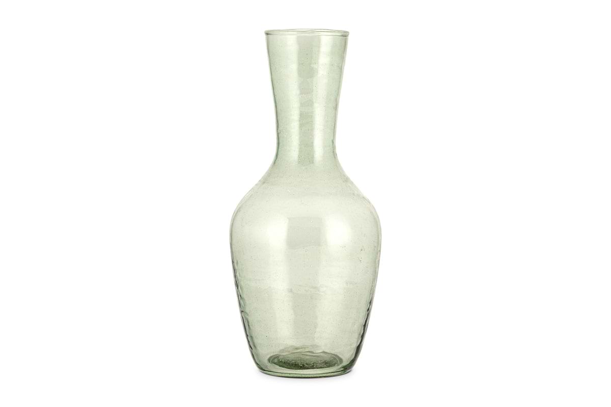The Yala Hammered Jug - Sage Green by nkuku is a tall, clear glass jug with a narrow neck and wider base, crafted from subtly textured, slightly green-tinted recycled glassware.