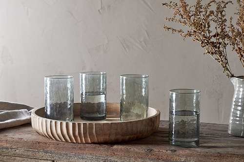 Four nkuku Yala Hammered Highball glasses in sage green sit on a rustic wooden table—three of the set of four are on a round wooden tray, with one beside it. A glass vase with dried branches is in the background against a light textured wall.