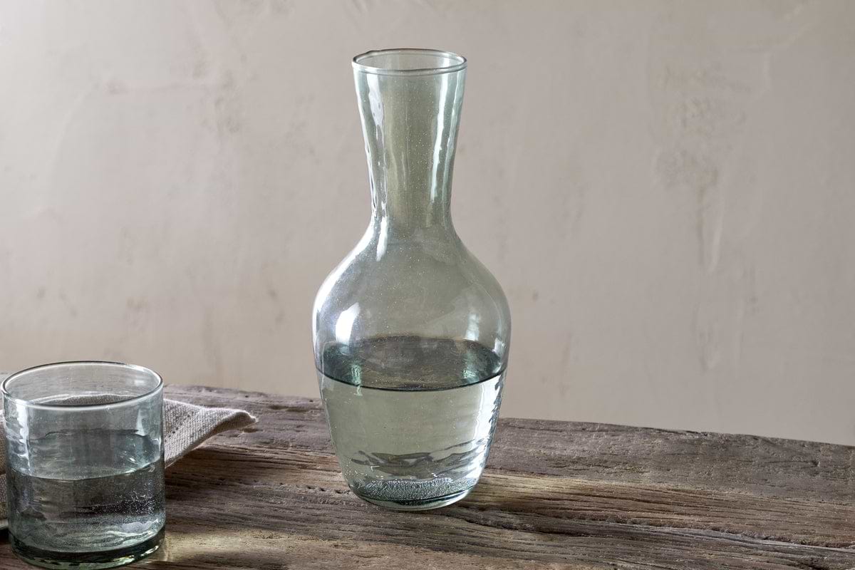 The nkuku Yala Hammered Jug in sage green, partially filled with water, sits on a rustic wooden table beside a glass tumbler and folded cloth, set against a neutral background.