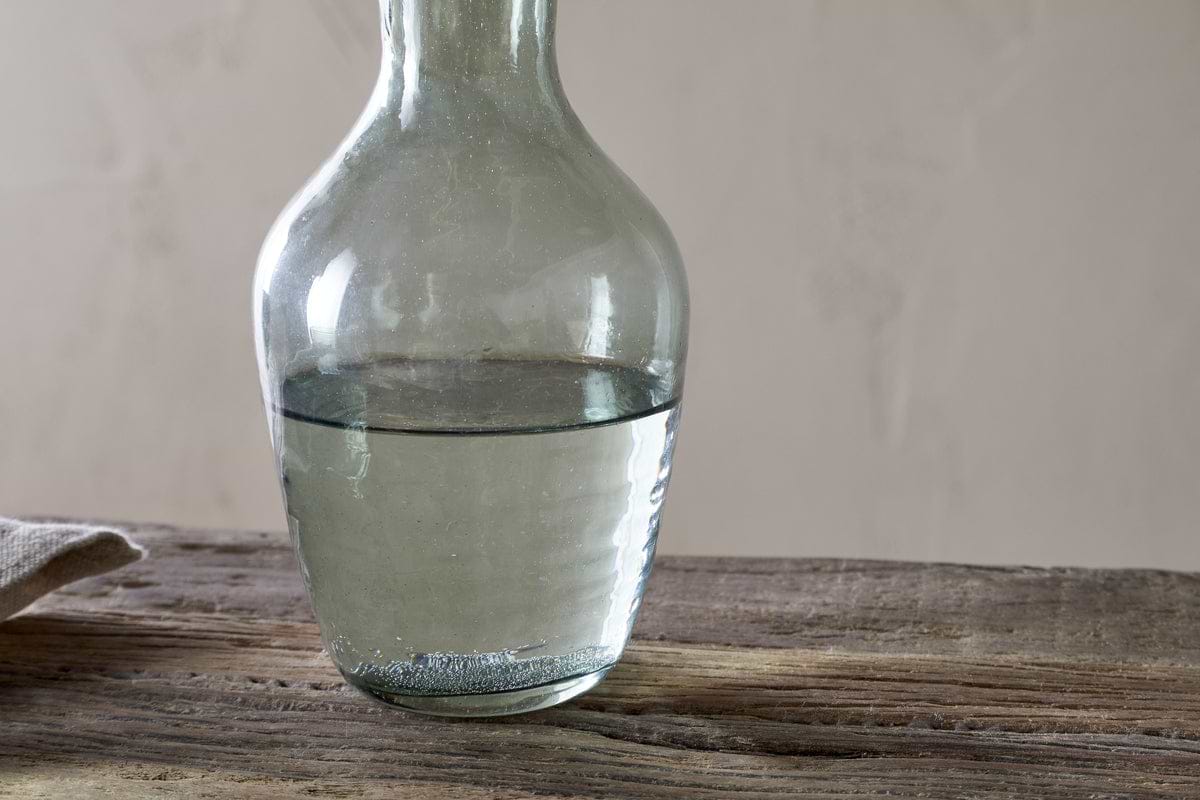 A nkuku Yala Hammered Jug in sage green, filled halfway with water, sits on a rustic wooden surface with a neutral beige background and a light-colored cloth partly visible to the left.