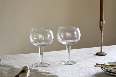Two nkuku Yala Hammered Gin Glasses (Set of 2) in clear, with textured design, rest on a light tablecloth next to a brass candlestick, gold utensils, and a folded napkin against a plain beige wall.