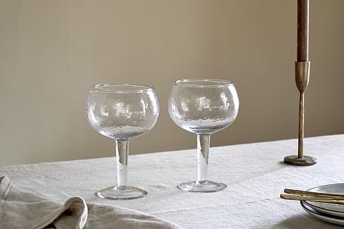 Two nkuku Yala Hammered Gin Glasses (Set of 2) in clear, with textured design, rest on a light tablecloth next to a brass candlestick, gold utensils, and a folded napkin against a plain beige wall.