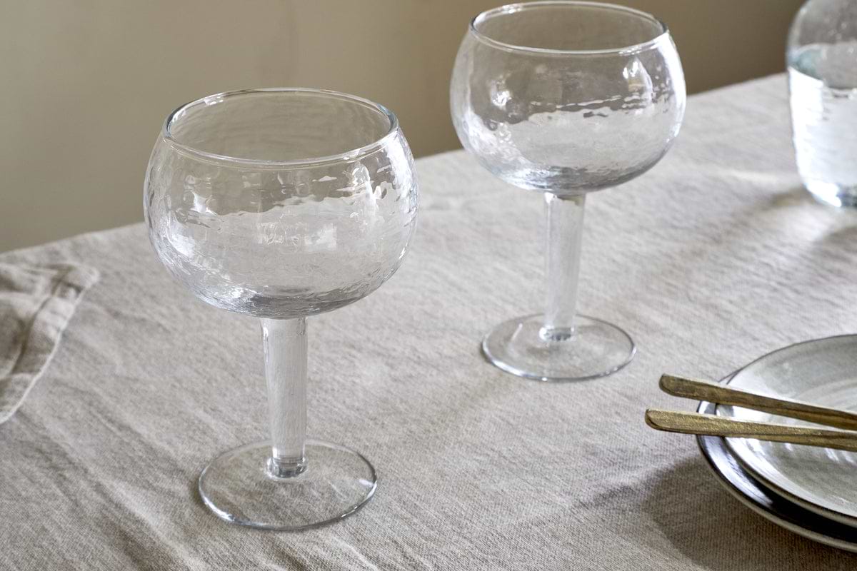 A set of two nkuku Yala Hammered Gin Glasses in clear, featuring a textured finish, rests on a beige tablecloth beside stacked plates with gold-toned cutlery and a glass of water in the background.