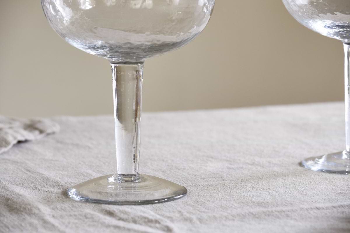 Close-up of the base and stem of two nkuku Yala Hammered Gin Glasses (Clear, Set of 2) on a light-textured tablecloth; a third goblet is partially visible on the right.