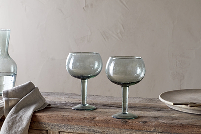 The nkuku Yala Hammered Gin Glass - Sage Green (Set of 2) features two round, clear glasses with green stems on a rustic wooden table, alongside a folded beige cloth, glass carafe, and plate against a textured neutral wall.
