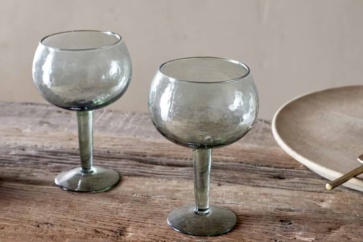 Two Yala Hammered Gin Glasses in sage green by nkuku, featuring short stems and a unique texture, are displayed on a rustic wooden table next to a beige plate.