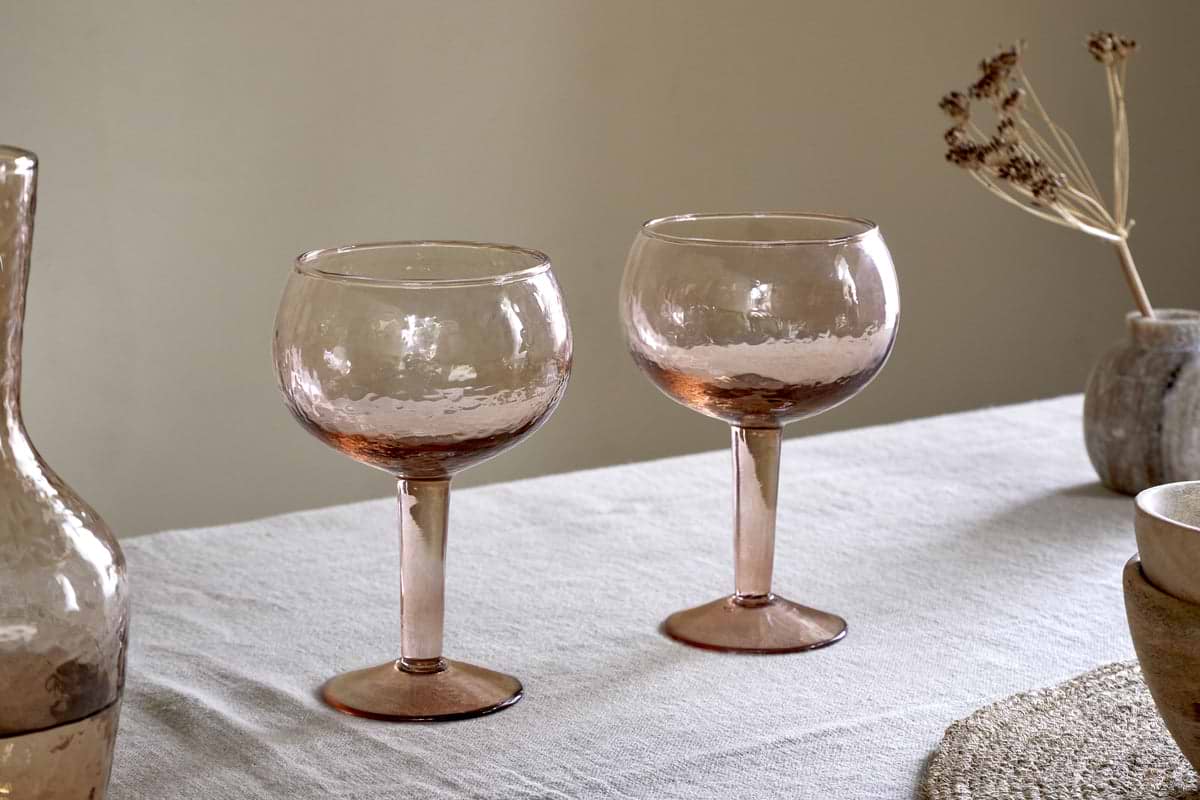 Two nkuku Yala Hammered Gin Glasses in Dusty Terracotta sit on a light tablecloth next to the matching pitcher and neutral ceramic bowls, with dried flowers in the background.