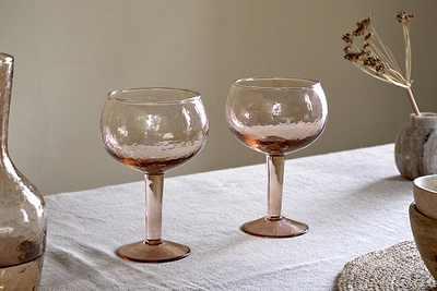Two nkuku Yala Hammered Gin Glasses in Dusty Terracotta sit on a light tablecloth next to the matching pitcher and neutral ceramic bowls, with dried flowers in the background.