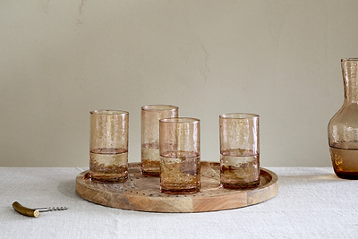Four nkuku Yala Hammered Highball glasses in Dusty Terracotta (set of 4) sit on a round wooden tray with water, next to a matching carafe and corkscrew, atop a white tablecloth against a light-colored background.