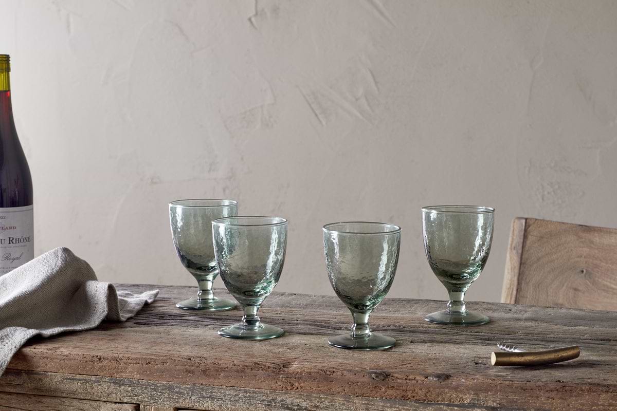 A set of four nkuku Yala Hammered Wine Glasses in Sage Green is arranged on a rustic wooden table with a folded linen napkin, corkscrew, and a partially seen wine bottle against a plain, light-colored wall.
