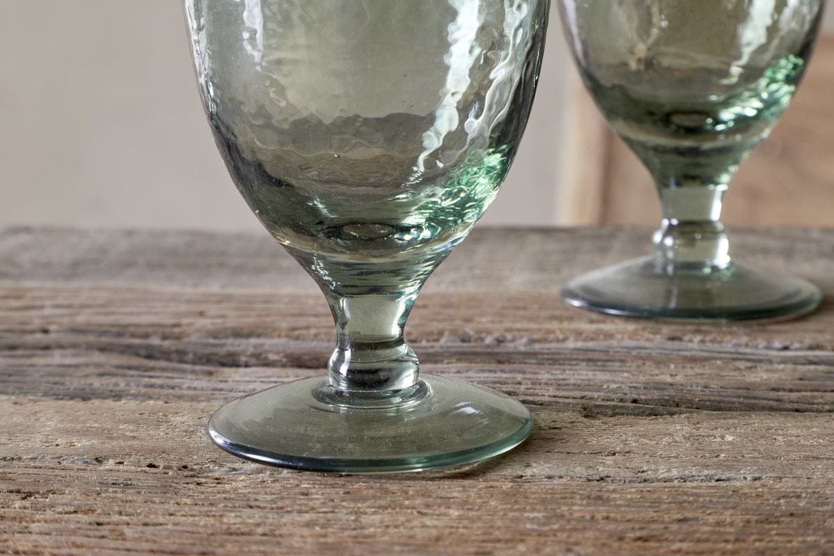 Close-up of the nkuku Yala Hammered Wine Glass in Sage Green (Set of 4), featuring thick stems on a rustic wooden surface, with one glass in sharp focus and another softly blurred in the background.