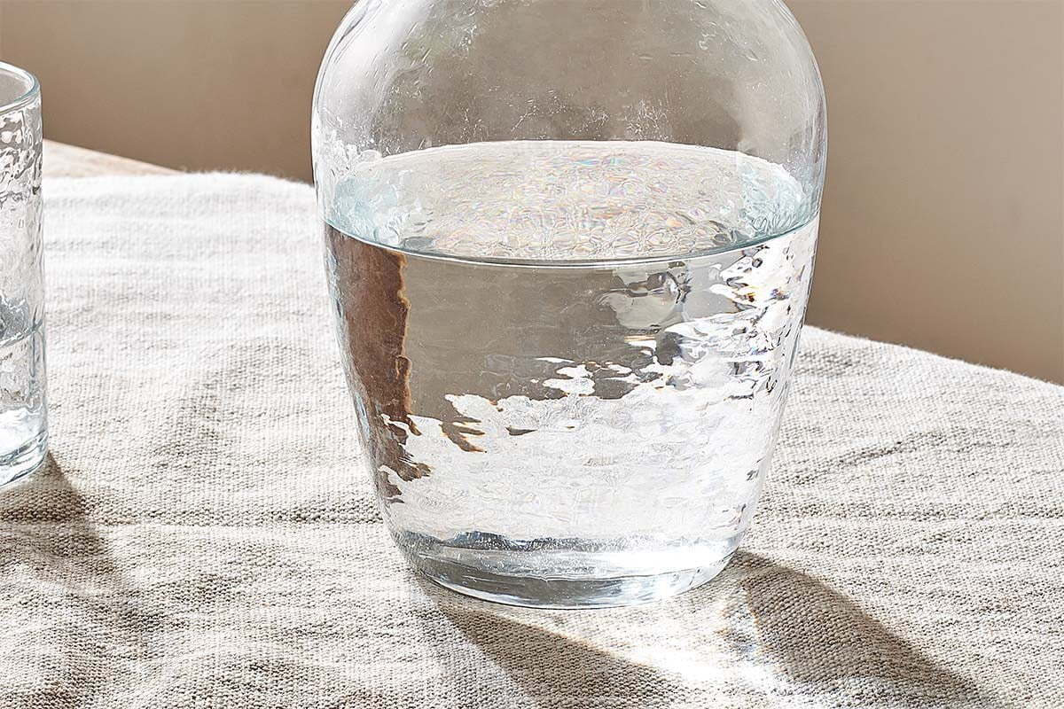 The nkuku Yala Hammered Jug - Clear Glass sits half-filled with water on a textured light tablecloth, sunlight reflecting off its hammered finish—a serene setting perfect for serving water or decanting wine.