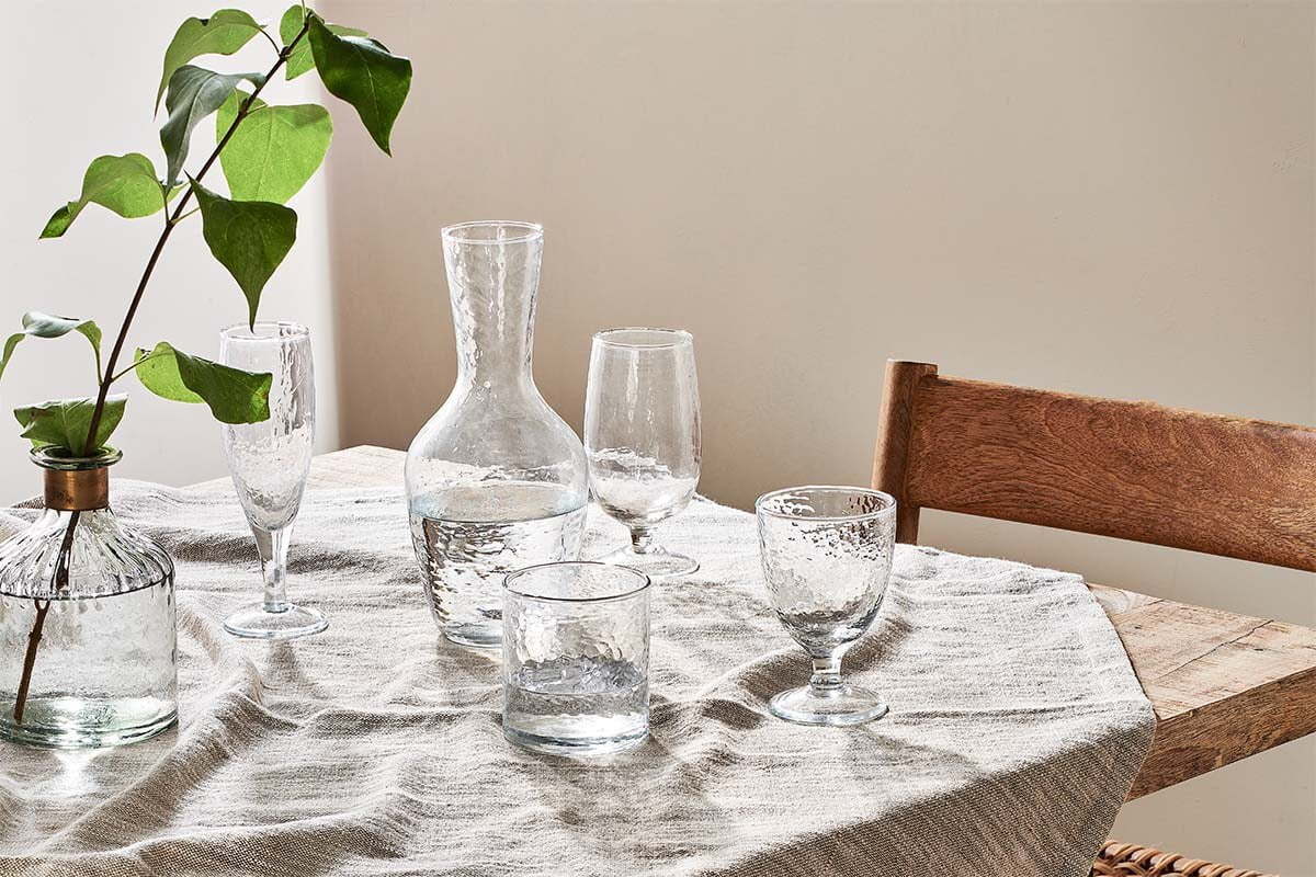 A wooden table with a beige textured tablecloth displays clear glassware—wine glasses, tumblers, an nkuku Yala Hammered Jug in clear glass, and a vase with green leafy stems. A wooden chair sits to the right. Sunlight creates a calm, elegant mood.