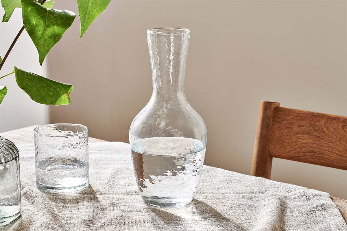 A nkuku Yala Hammered Jug in clear glass, half-filled with water, sits on a textured tablecloth beside a matching glass and green-leaf vase. A wooden chair is partly visible to the right against a plain, light wall.