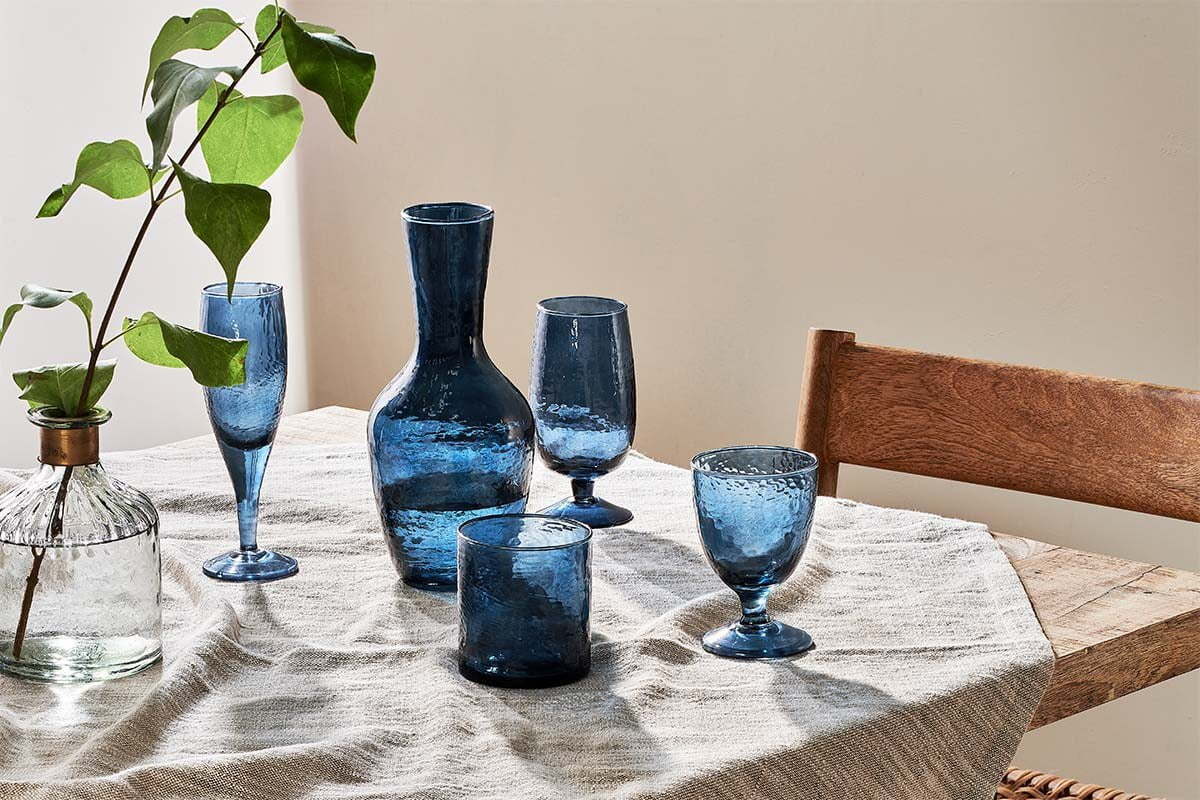 A wooden table with a beige cloth displays four nkuku Yala Hammered Tumblers in indigo. To the left is a textured glass bottle with green stems; a wooden chair appears partly on the right.