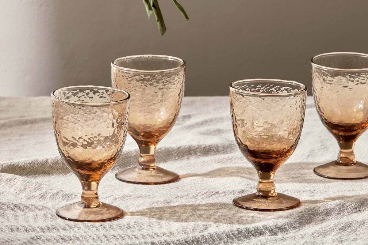 A set of four nkuku Yala Hammered Wine Glasses in Dusty Terracotta sit atop a white, slightly wrinkled tablecloth, sunlight enhancing their translucent, textured glass. A small green leafy stem hangs from the top edge.