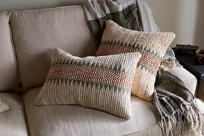 Two nkuku Zairya Jute & Cotton Cushion Covers in Natural & Rust add warmth to a light beige couch. A textured neutral throw completes the cozy look, while a small table sits beside the sofa.
