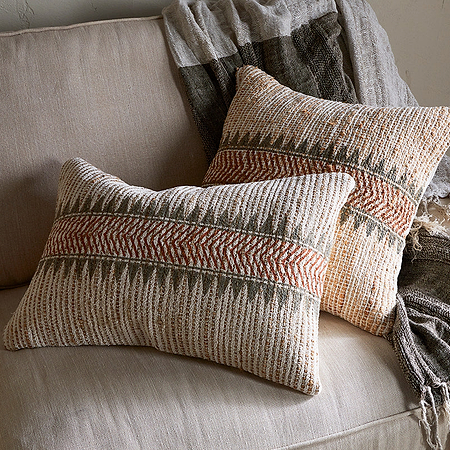 Two nkuku Zairya Jute & Cotton Cushion Covers in Natural & Rust add warmth to a light beige couch. A textured neutral throw completes the cozy look, while a small table sits beside the sofa.