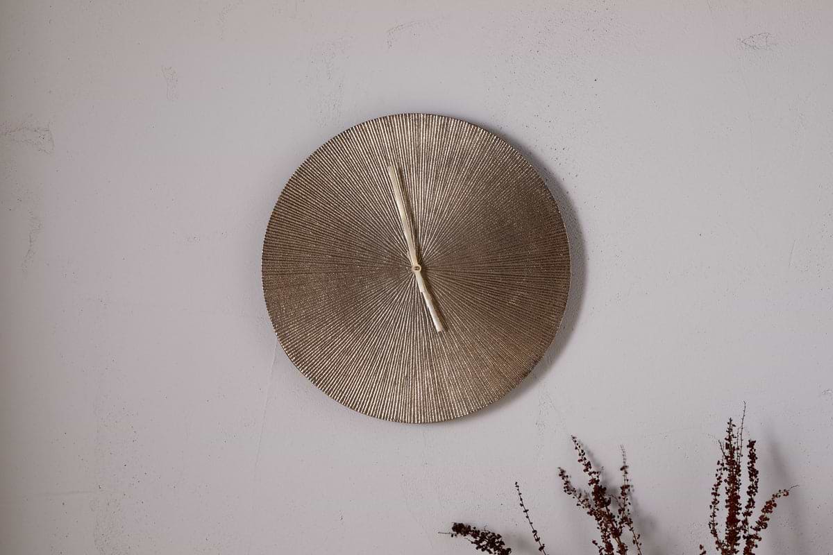 A round, textured Zuwena Etched Wall Clock in antique brass by nkuku, with no numerals and hands at 12:00, hangs on a light gray wall. Dried brown foliage in the corner adds a natural touch to this minimalist display.