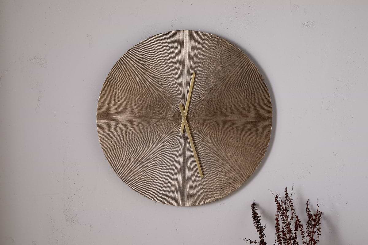 The nkuku Zuwena Etched Wall Clock in Antique Brass, featuring a textured radial gold face and minimal gold hands, hangs on a plain light wall with dried brown plant stems in the corner for a minimalist accent.