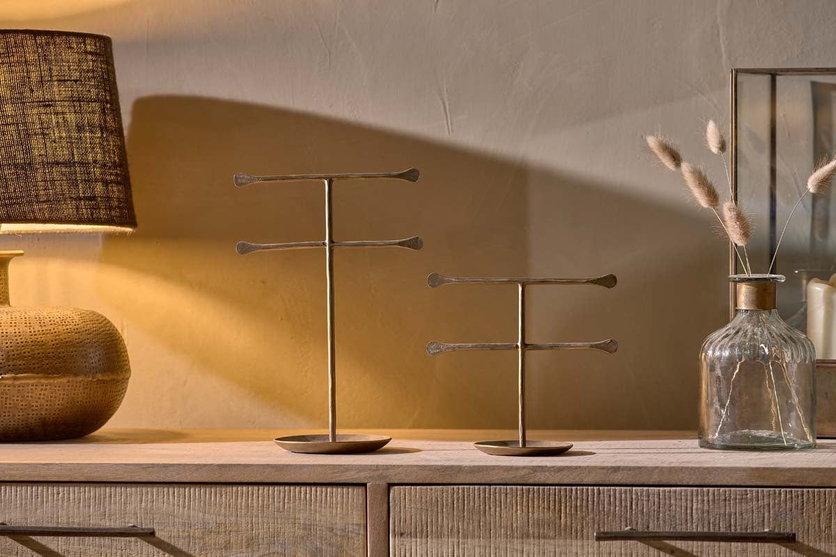 Two nkuku Liman Jewellery Stands with horizontal brass bars are displayed on a wooden dresser. A tan lamp with a textured shade sits to the left; to the right, a clear glass vase holds dried grass, all softly lit for a cozy, warm mood.