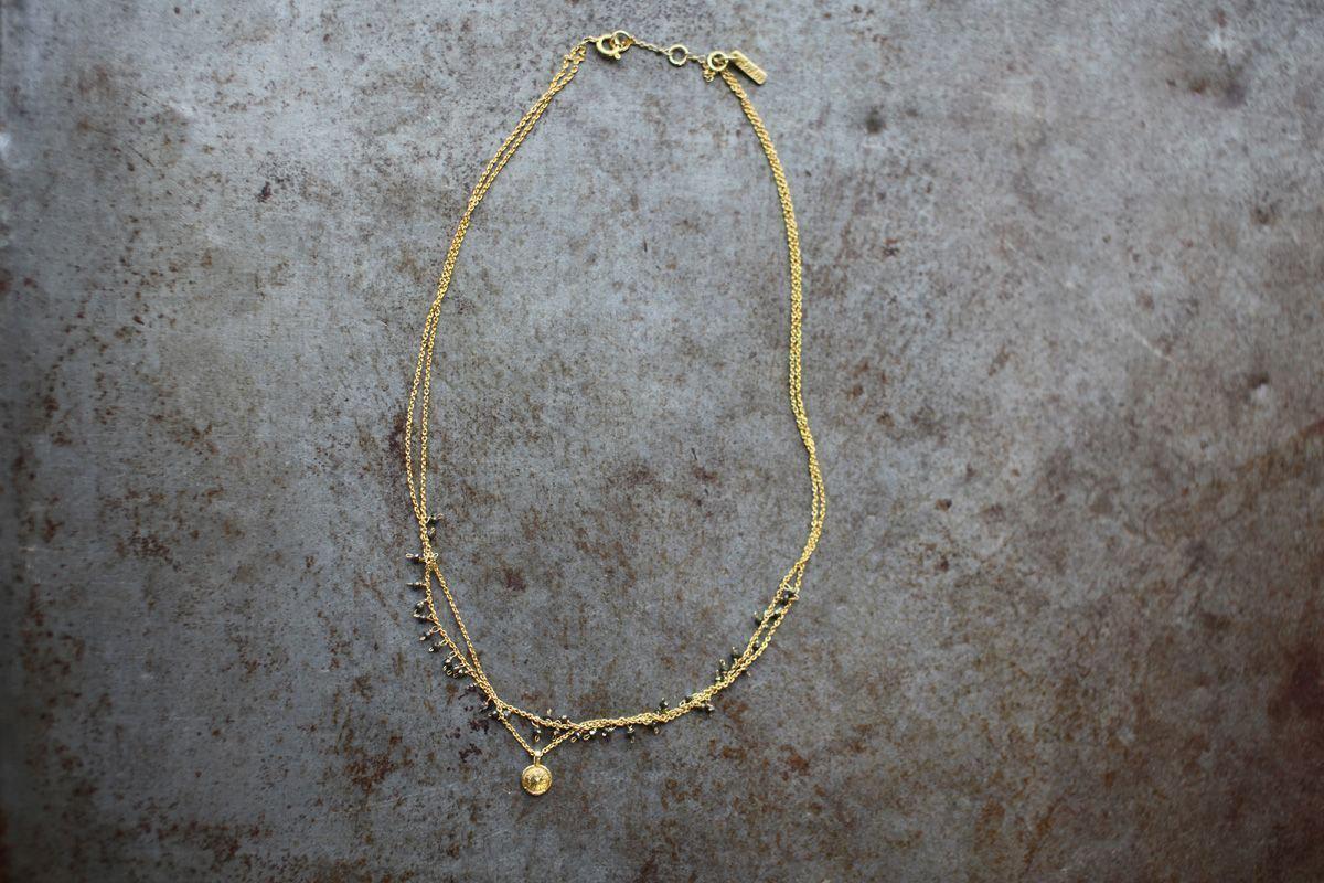 The Mai Necklace by nkuku features a delicate, gold-toned asymmetric design with two fine chains—one with small beads and the other with a round pendant—plus a lobster clasp and branded tag near the closure. Photographed on textured gray.