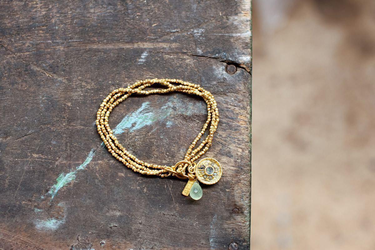 The nkuku Pandita Charm Bracelet features a gold-plated design with three charms—a coin, a rectangular bar, and a light green teardrop—resting on a rustic wooden surface. Ideal for fans of handmade jewelry.