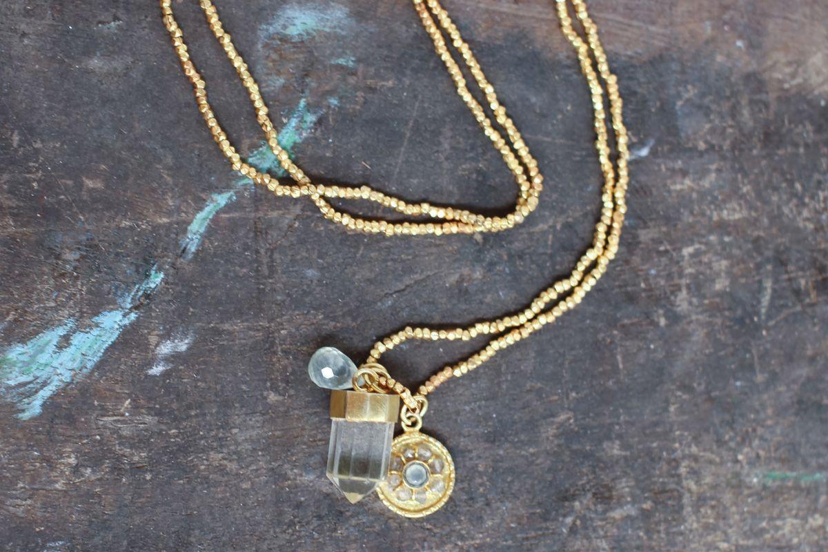 The Pandita Charm Necklace by nkuku rests on a rustic wooden surface, featuring three pendants: a crystal quartz point, a clear round stone, and a gold floral medallion with a gemstone. This elegant piece is handmade.